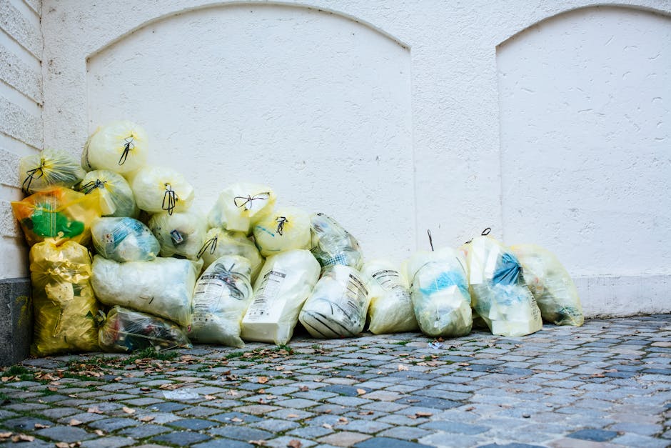 A collection of yellow and white trash bags filled with waste material, piled against a white exterior wall on a cobblestone street. The bags appear to contain household rubbish such as paper, plastic