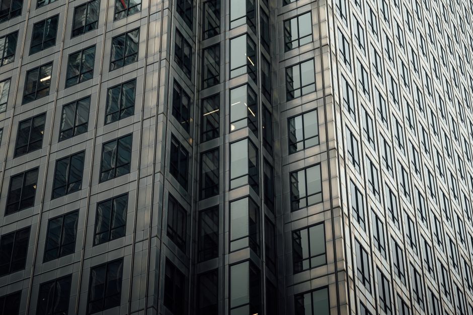 Close-up view of a modern commercial office building with a glass and metal facade, featuring numerous reflective windows arranged in a grid pattern. The building's exterior surface appears clean and