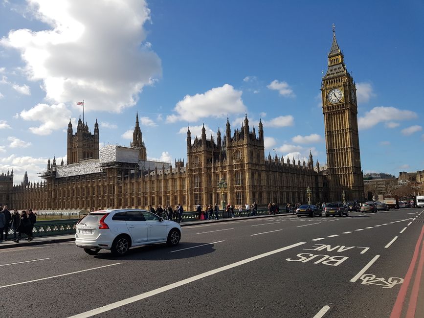 A view of Westminster Bridge in London with several vehicles including a white van and a blue bus traveling across. In the background, the iconic Big Ben clock tower and the Houses of Parliament are v